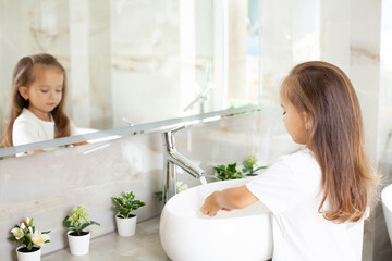 A beautiful cute little girl washes her hands in the bright bathroom in front of the mirror....