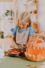 a blonde baby girl with long hair in an apron in the kitchen, decorated with pumpkins and garlands for Halloween, prepares a focaccia pie. space for text. High quality photo