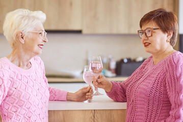 two stylish senior women in pink sweaters drinking rose wine at modern kitchen gossiping. Friendship, talk, gossip, event, relationships, news, family concept 
