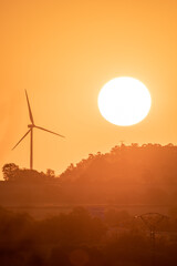 Vertical image of Sunrise with a large sun rising from behind a mountain next to a renewable energy wind turbine during the golden hour.