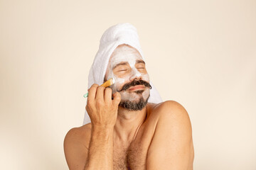Happy man at facial spa day puts on facial mask. Adult male with towel on head, beard and mustache.