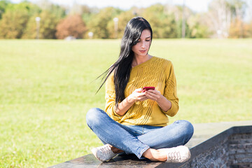 Obraz premium A beautiful and attractive Caucasian brunette girl sitting and using a smartphone in the park.