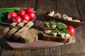 On a wooden background, there are ready-made sandwiches with cod liver, tomato and greens, canned fish are lined in a dark plate. 