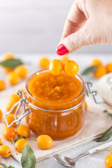 Woman hand holds yellow cherry plum over glass jar of homemade DIY natural canned yellow cherry plum sauce chutney with chilli or tkemali standing on white table