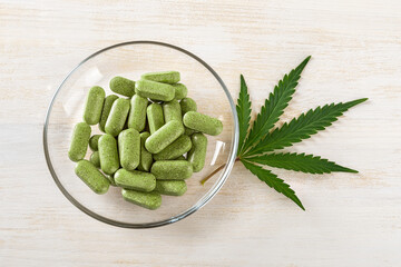 Prescription cbd pills of cannabis sativa weed on a glass saucer and fresh green marijuana leaf over white wooden table. Medical hemp pills. Marijuana plant for herbal alternative medicine.