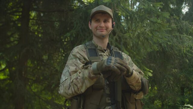 Portrait of cheerful young adult soldier in camouflage with tactical headset looking directly at camera, smiling outdoors. Joyful military man simulating radio call, using walkie-talkie in forest area