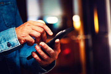 Primer plano de la mano de un hombre sosteniendo su smartphone. Enviando un sms y compartiendo contenidos en las redes sociales desde el interior de la vivienda - obrazy, fototapety, plakaty