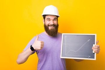 Bearded male engineer is wearing hard hat while holding thumb up and a chalkboard with a graphic over yellow background.