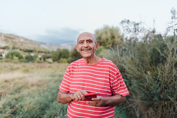 Middle-aged man looking at the camera and smiling while using a mobile phone outdoors in the...