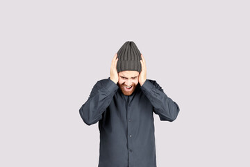 Young Latino man in shirt and knit hat screaming, stressed, covering his ears with his hands, does not want or avoids hearing bad news, isolated on gray background. Copy space.