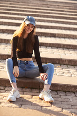 Beautiful smiling teen woman in blue jeans and cap looking happy sitting street stairs on summer  background. Closeup portrait