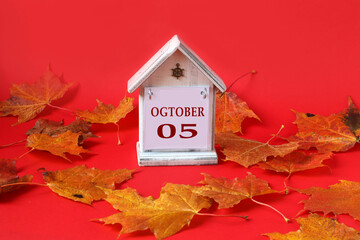 Calendar for October 5: decorative house with the name of the month in English, the numbers 05, yellow autumn maple leaves scattered on a red background
