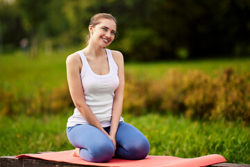 Smiling woman sits on exercising mat during yoga in city garden.
