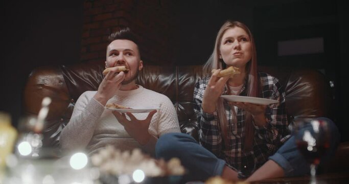 Surprised Millennial Couple Sitting On Couch Watching TV Show Or Movie At Home In Evening. Handsome Man And Attractive Woman Eating Pizza Shocked By News They Saw. Wide Angle Dolly Shot