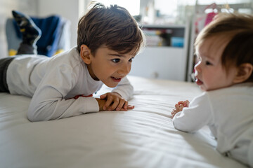 Small caucasian boy taking care of his baby brother or sister while while playing on the bed at home in day brotherhood and family growing up concept