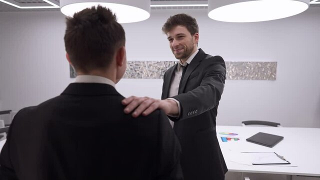 Positive handsome man tapping shoulder of colleague endorsing coworker in office indoors. Portrait of satisfied manager supporting employee talking and smiling. Slow motion