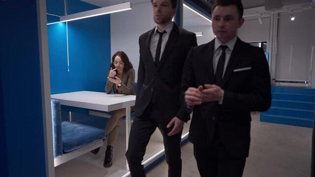 Confident Caucasian businessmen passing in corridor with absorbed businesswoman sitting in glass office surfing Internet on smartphone. Busy employees indoors at workplace. Slow motion