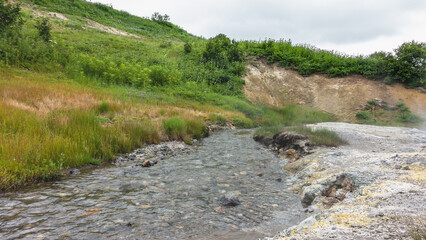 A clear stream flows from hot springs along a rocky bed. There are sulfur deposits on the shore. Steam and haze above the ground. There is green vegetation on the hillside. Kamchatka