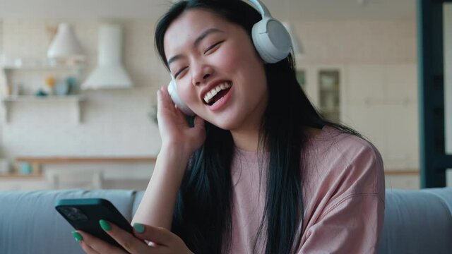 Attractive woman of Asian ethnicity wearing headphonees, singing, dancing and listening to the music, enjoying her day off, holiday at home. Time to have a rest and fun. Home leisure activity concept