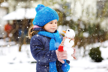 Cute little toddler girl making mini snowman and eating carrot nose. Adorable healthy happy child playing and having fun with snow, outdoors on cold day. Active leisure with children in winter