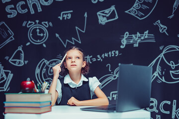 Serious thoughtful schoolgirl raising pen up. Sitting at the desk with laptop. Distance education 