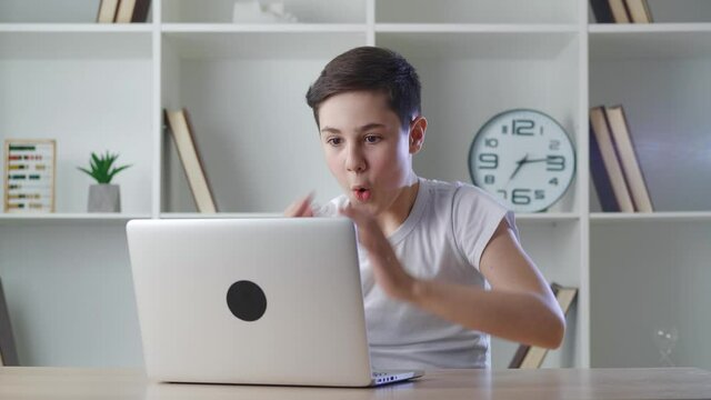 A shocked 13-year-old boy is frightened by what he sees on a laptop screen sitting at a table at home. Astonished teenager surprised by watching content on computer.