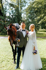 Happy beautiful young bride and groom hugging next to horse and looking at camera, married...