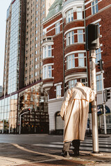 Vertical shot of an old man in a beige cloak leaning on the pole in the city with highrise buildings