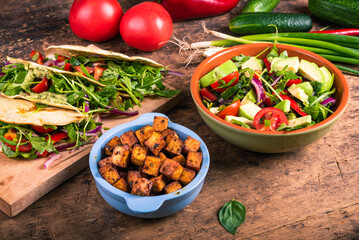 Fried diced tofu cheese in blue bowl, bowl of vegan salad and tacos with vegetable salad on wooden board and ingredients - vegetarian cuisine.