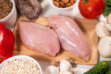 Raw chicken breast fillet on a wooden cutting board surrounded by various vegetables and other foods. The process of preparing a delicious dinner. Close-up, top view.