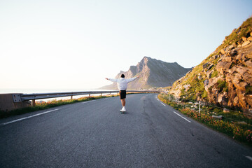 Wide shot of man in oversized hoodie and shorts ride on longboard into mountain road landscape on...