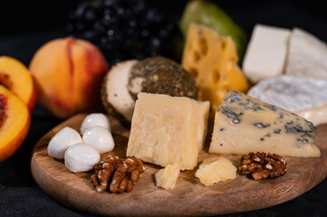 Various types of cheese on a dark background. Nearby are peaches, grapes, nuts. An exquisite delicacy. Close-up.