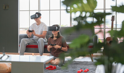 Young attractive Asian couple holding joysticks  enjoy playing video games together wearing VR glass with fun and excitement in living room with glasses windows background