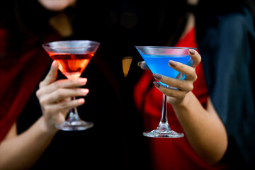 Unrecognizable sexy female in black and red witch dress costume tall hat and shawl holding glass of cocktail cheers and happy together on light bokeh background in Halloween party
