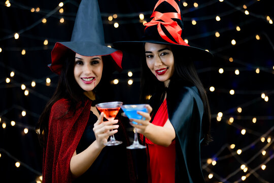 Caucasian and Asian sexy female in black and red witch dress costume tall hat and shawl holding glass of cocktail cheers and happy together on light bokeh background in Halloween party