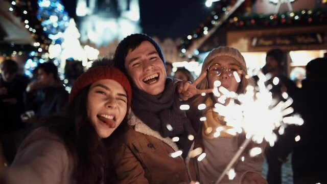 Film Shot. Prores 422. Three happy friends with sparklers call their relatives via video link on new year's eve to the sound of chimes in the middle of the street in the lights of the Christmas market