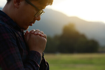 Religious man praying to God resting his chin on his hands in a field during a beautiful sunset.His...