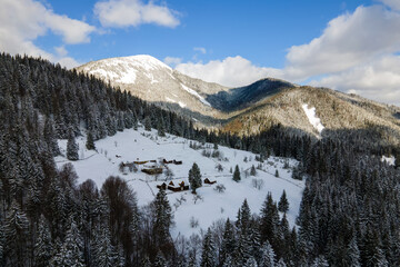 Aerial winter landscape with small rural houses between snow covered forest in cold mountains.