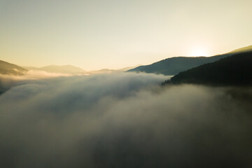 Aerial view of vibrant sunrise over white dense fog with distant dark silhouettes of mountain hills on horizon.