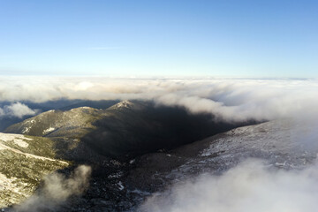 Aerial view from above of white puffy clouds covering snowy mountain tops in bright sunny day.