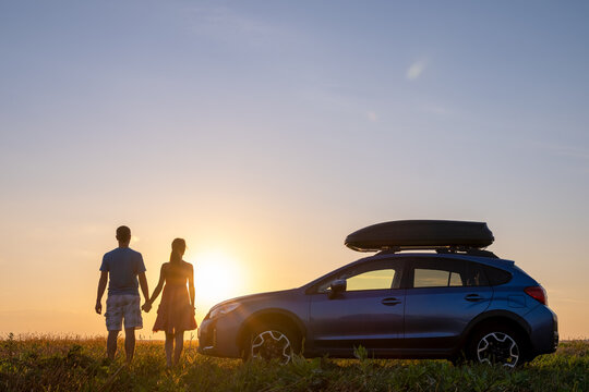 Happy couple relaxing beside their SUV car during honeymoon road trip at sunset. Young man and woman enjoying time together travelling by vehicle.