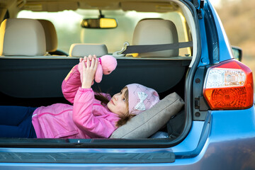 Pretty happy child girl playing with a pink toy teddy bear in a car trunk.