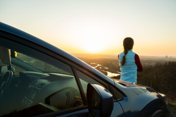 Young woman standing near her car enjoying warm sunset view. Girl traveler leaning on vehicle hood looking at evening horizon.