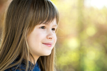 Fototapeta premium Portrait of pretty child girl with gray eyes and long fair hair smiling outdoors on blurred bright background. Cute female kid on warm summer day outside.