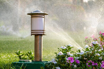 Plastic sprinkler irrigating flower bed on grass lawn with water in summer garden. Watering green vegetation duging dry season for maintaining it fresh.