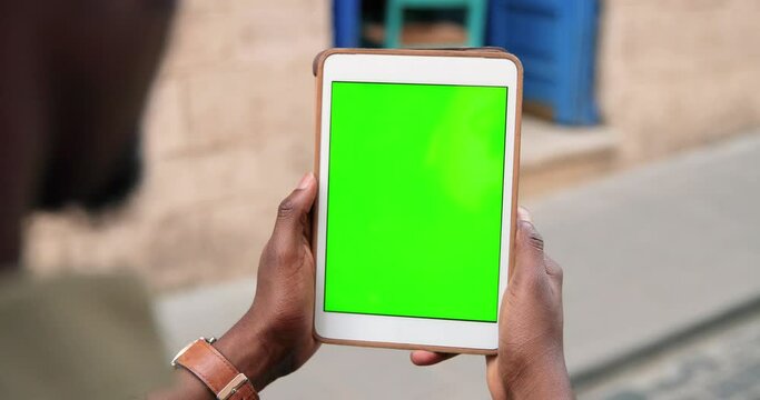 Back view of multiracial guy standing at the street and scrolling at the tablet computer. Rear of man working outdoors. Green screen and chroma key concept