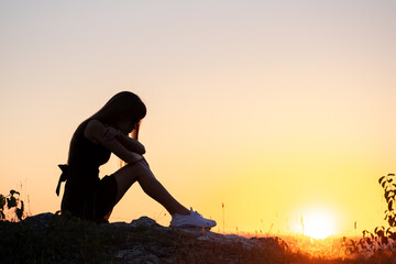 Pretty sad woman in black short summer dress sitting on a rock thinking outdoors at sunset. Fashionable female contemplating in warm evening in nature.
