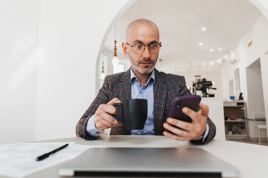 Surprised businessman looking at mobile phone and holding a mug with coffee - Powered by Adobe