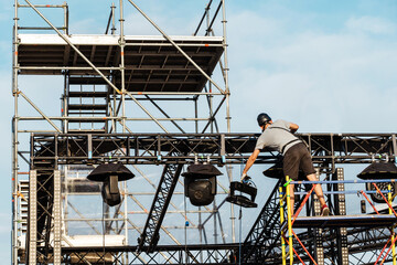 Installation of lighting equipment on the concert stage. A worker installs a powerful lantern on a metal frame under the open sky. Preparation for mass events. Unrecognizable person