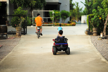 Obraz premium Rear view of boy drives the electrical car toy on the road inside the house with blurred people cycling the bicycle in background. Quarantine at home